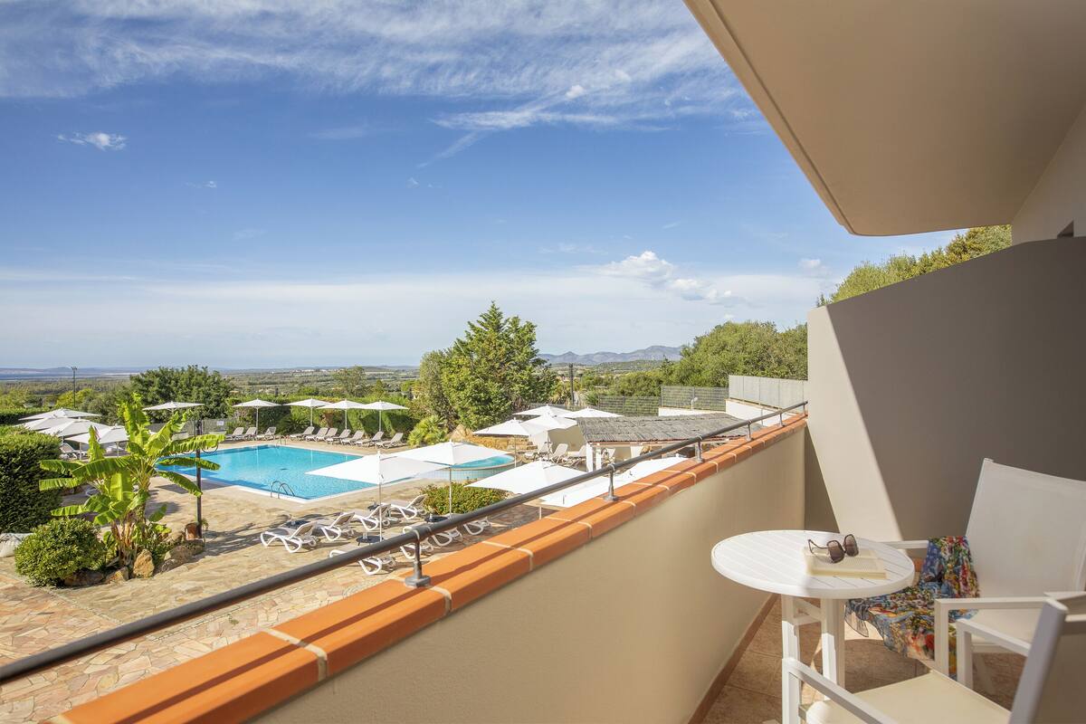Ein Balkon mit Blick auf einen Pool, weiße Liegestühle und ein Tisch mit Sonnenbrillen.