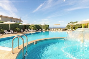 A swimming pool with lounge chairs and umbrellas, surrounded by greenery and a building.