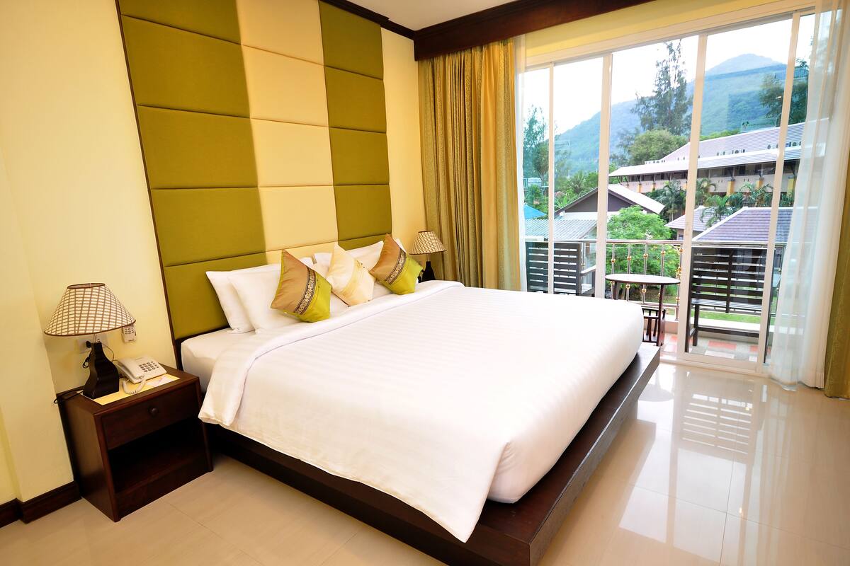 A hotel room with a large bed, a nightstand with a lamp, and a view of the outdoors.