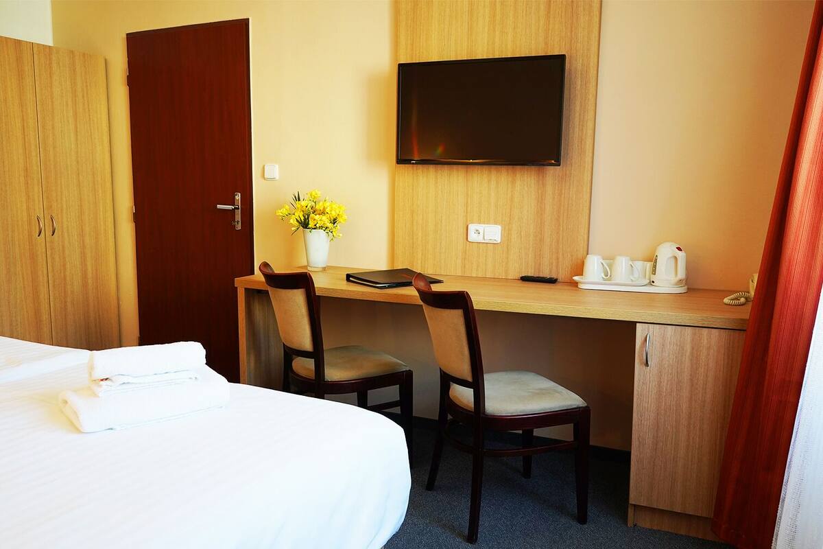 A hotel room with a bed, a desk with two chairs, a television, a vase with flowers, and a coffee maker.