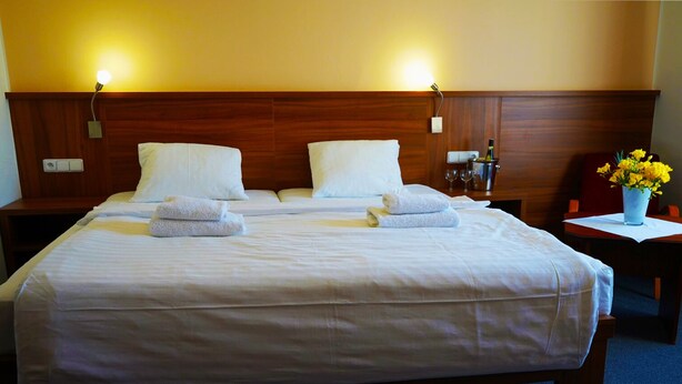 Cama doble con sábanas y toallas blancas, cabecera de madera, mesitas de noche y un florero con flores amarillas.