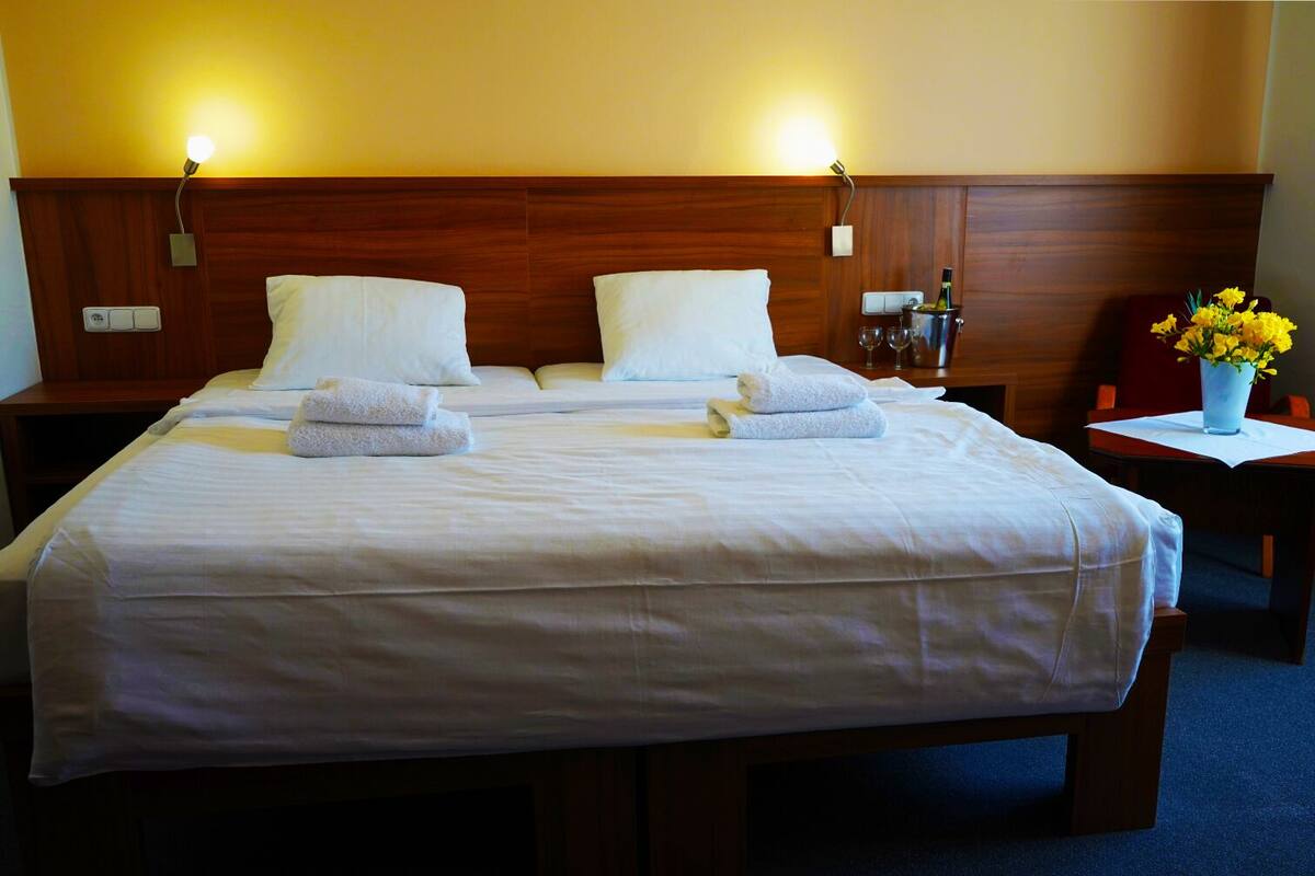 A double bed with white linens and towels, wooden headboard, bedside tables, and a vase with yellow flowers.
