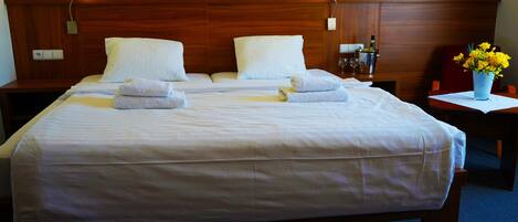 Cama doble con sábanas y toallas blancas, cabecera de madera, mesitas de noche y un florero con flores amarillas.