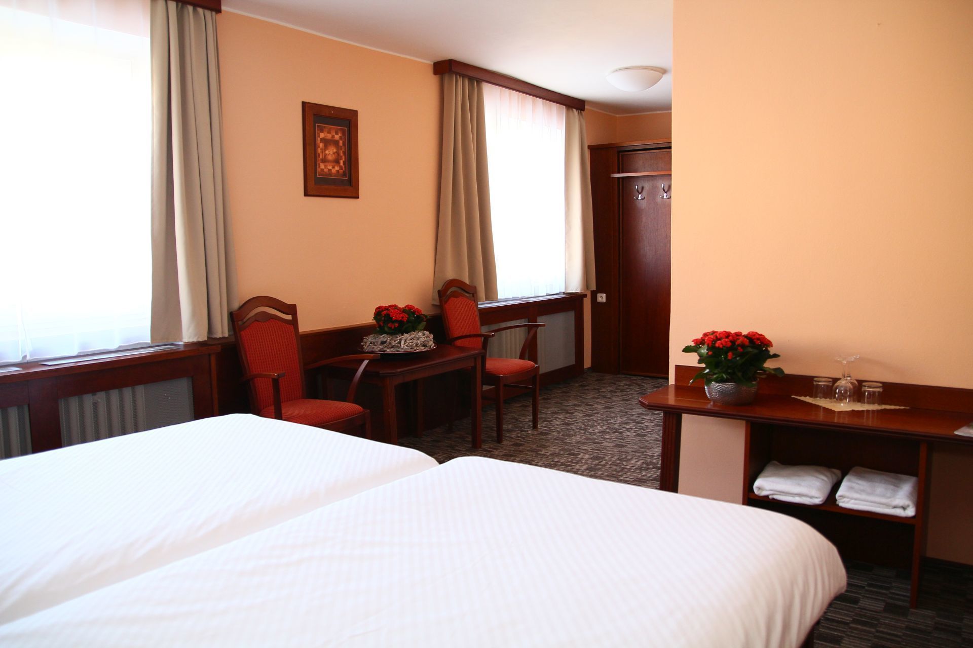 A hotel room with two beds, a desk with a chair, a small table with a vase of flowers, and a door.