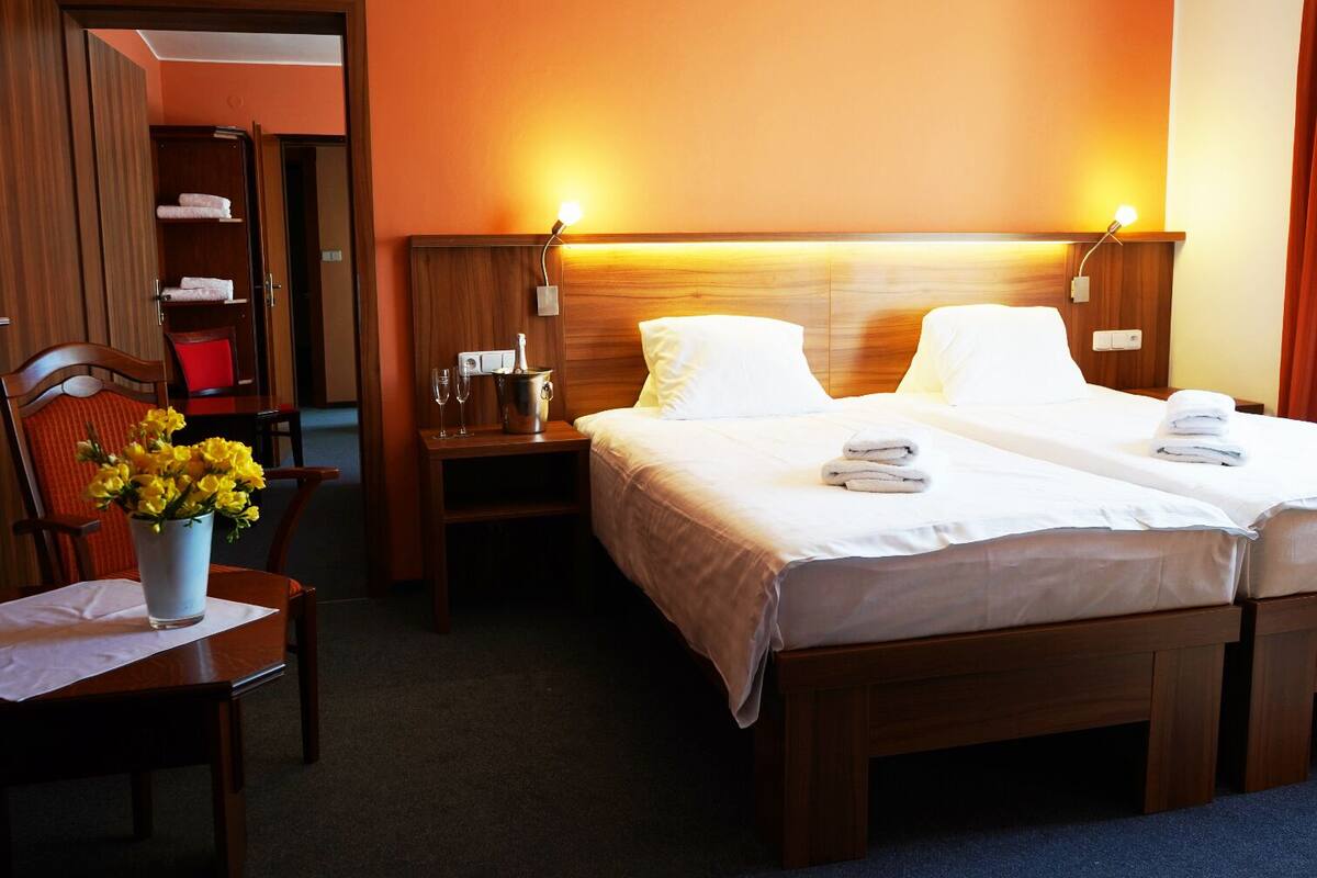 A hotel room with two beds, a wooden headboard, bedside lamps, a nightstand with a bottle and glasses, a chair with a vase of flowers, and a wooden wardrobe.