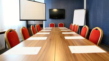 A conference room with a long wooden table, red upholstered chairs, a large screen, and a whiteboard.