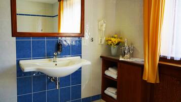 Un baño con un lavamanos blanco, azulejos azules, un mueble de tocador de madera, un espejo y un florero con flores amarillas.