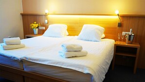 Habitación de hotel con cama doble, dos almohadas blancas, cabecera de madera, mesita de noche con un jarrón de flores amarillas y un minibar.