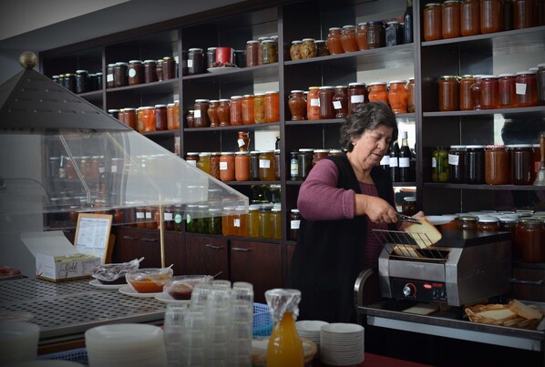 A kitchen area with a woman preparing food, shelves stocked with jars, and a menu stand. - Elyssia Hotel (Pedhoulas)