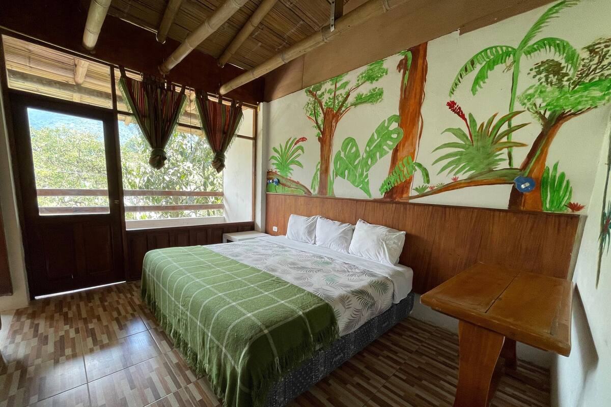 Una habitación con una cama, cabecera de madera, ventana con cortinas y una pared decorada con un mural de temática natural, además de una mesa de madera.