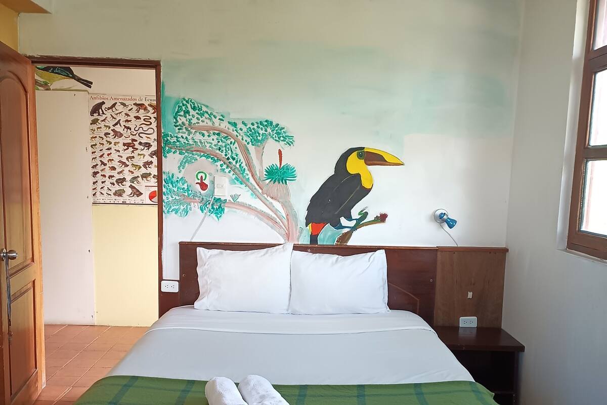 Una habitación con una cama, un edredón verde a cuadros, dos toallas enrolladas, un cuadro en la pared de un tucán y una puerta con un cartel.