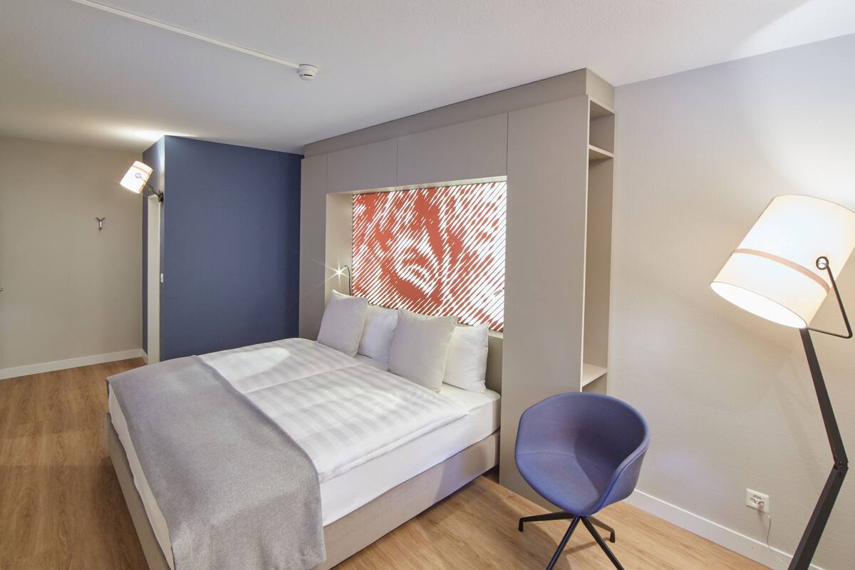 A modern bedroom with a bed, a chair, a lamp, and a wall with a red pattern.