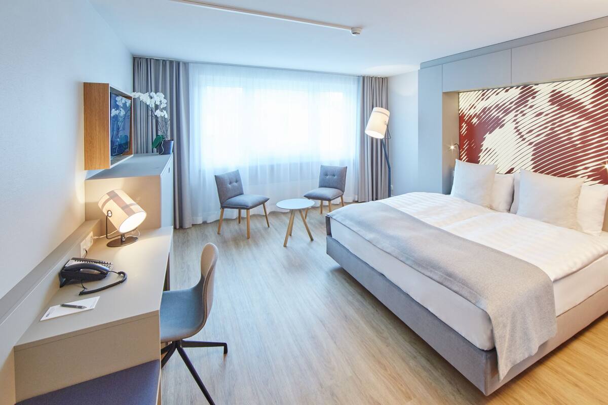 A modern hotel room with a large bed, a desk with a lamp, two chairs, a small table, and a large window with curtains.