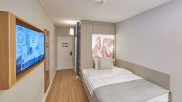 A modern hotel room with a large flat-screen TV mounted on the wall, a bed with white bedding, a wooden floor, and a window with blinds.