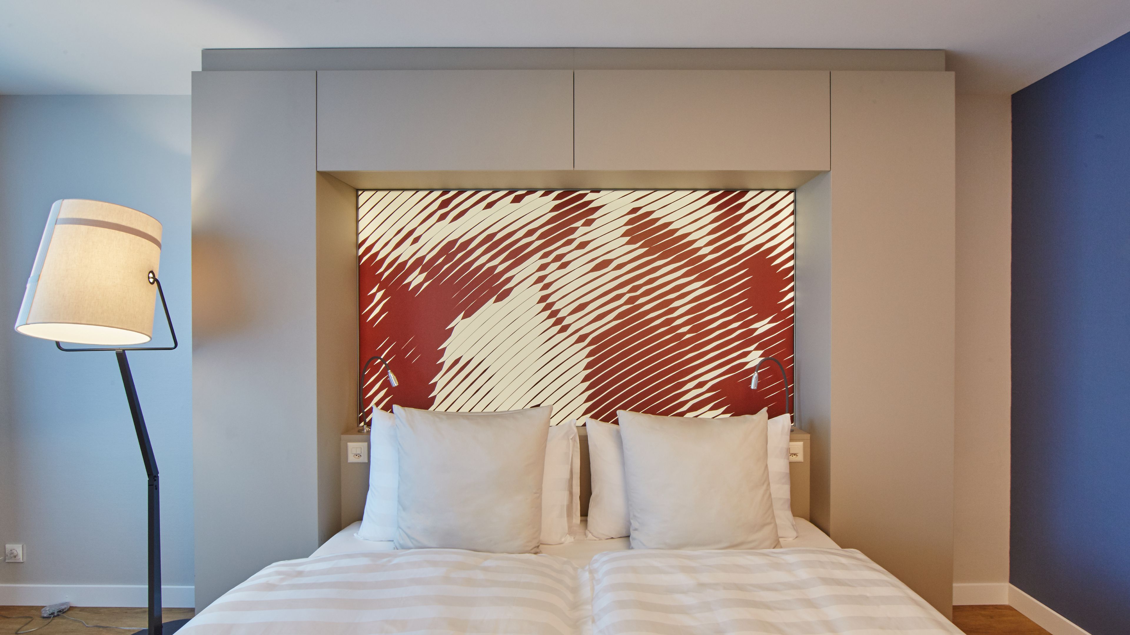 A modern hotel room with a large bed, two pillows, a headboard with a red and white abstract design, and a floor lamp.