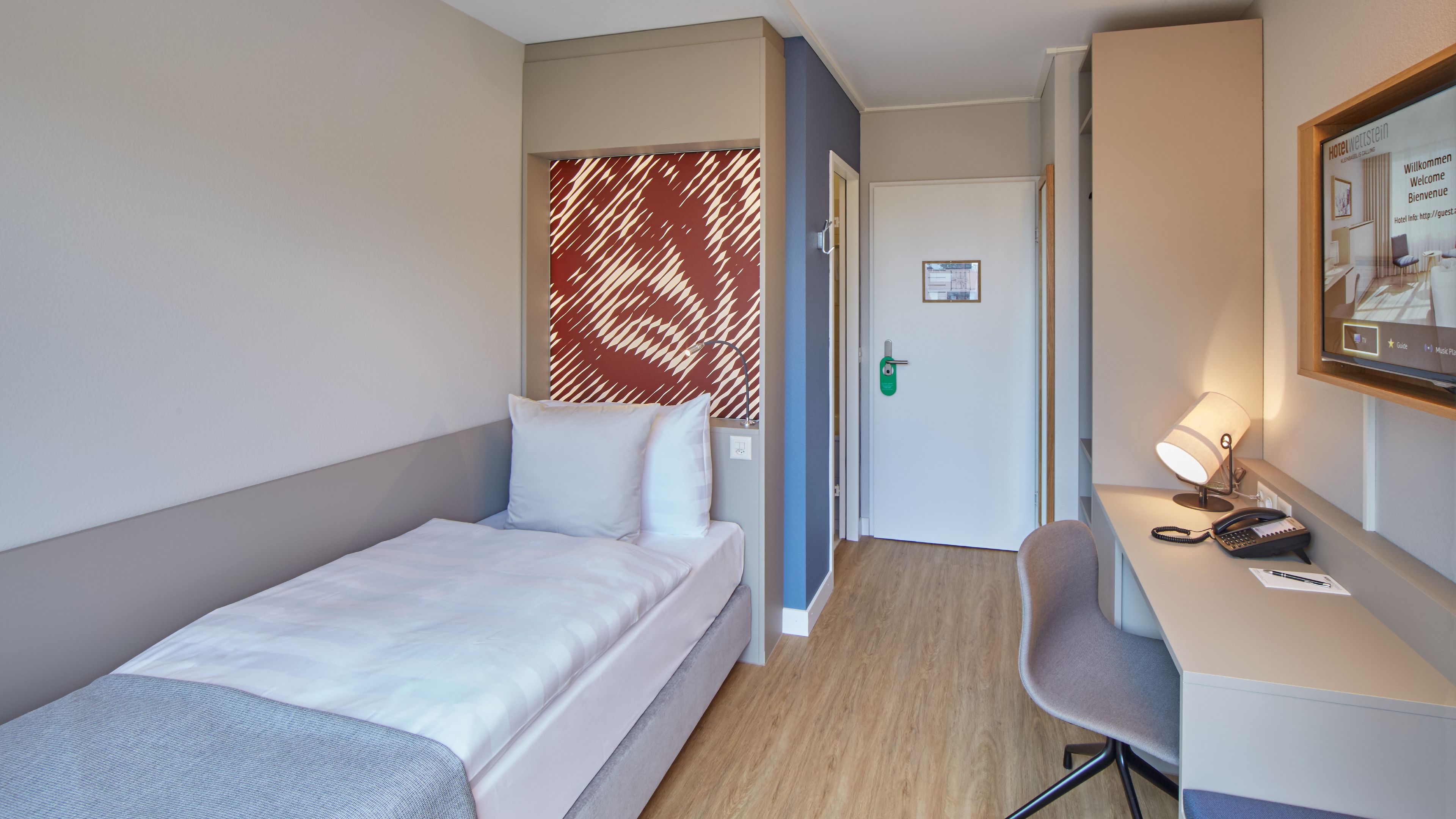 Habitación de hotel con una cama individual, un escritorio con silla, una lámpara de escritorio, un teléfono y un cartel de bienvenida enmarcado.