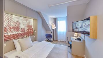 A modern hotel room with a bed, a desk, a chair, a TV, and a window with blinds.