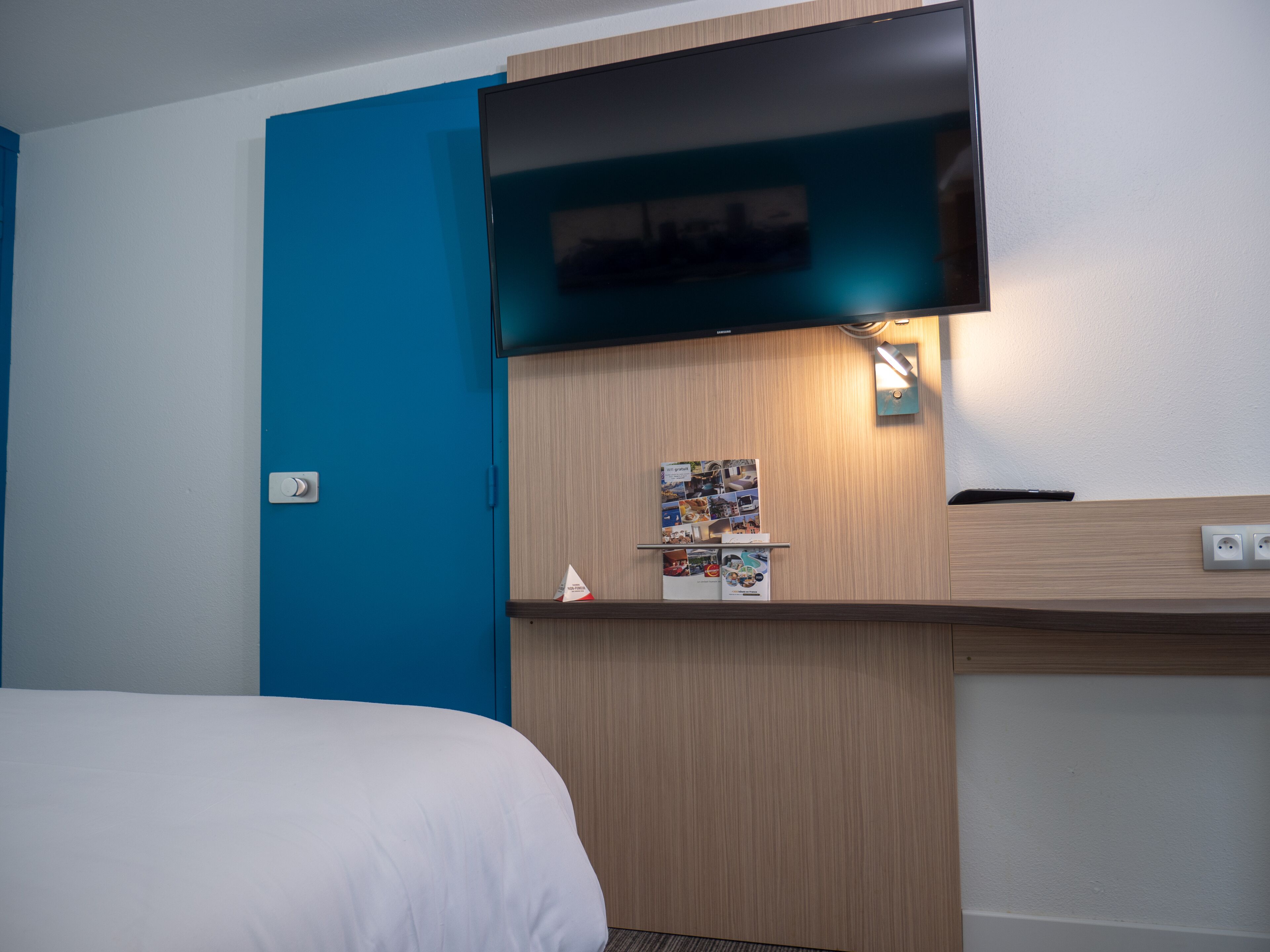 A hotel room with a bed, a flat-screen TV mounted on a wooden panel, a wall-mounted light, and a small shelf with brochures.