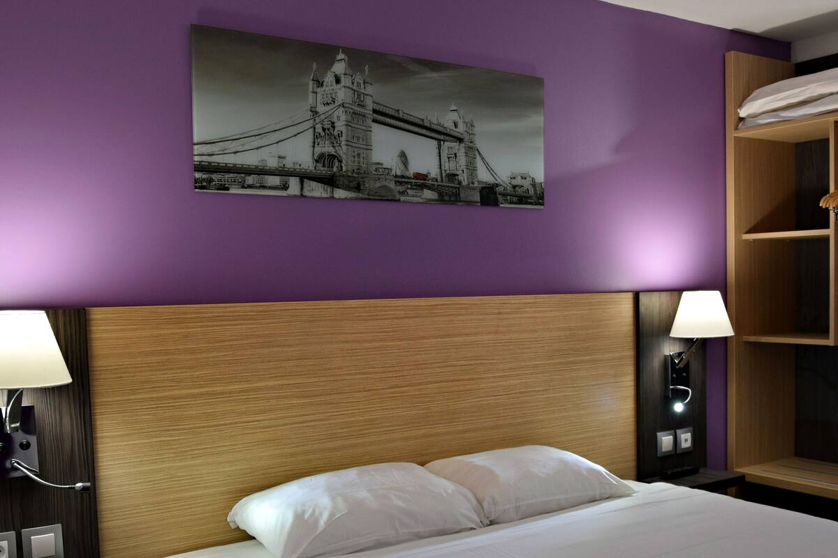 A hotel room with a bed, two bedside lamps, a wooden headboard, a wall-mounted shelf with a towel, and a picture of a bridge.
