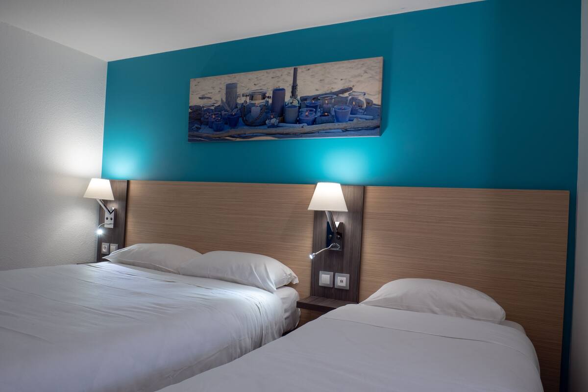 A hotel room with two beds, a cityscape painting on the wall, and two wall-mounted lamps.