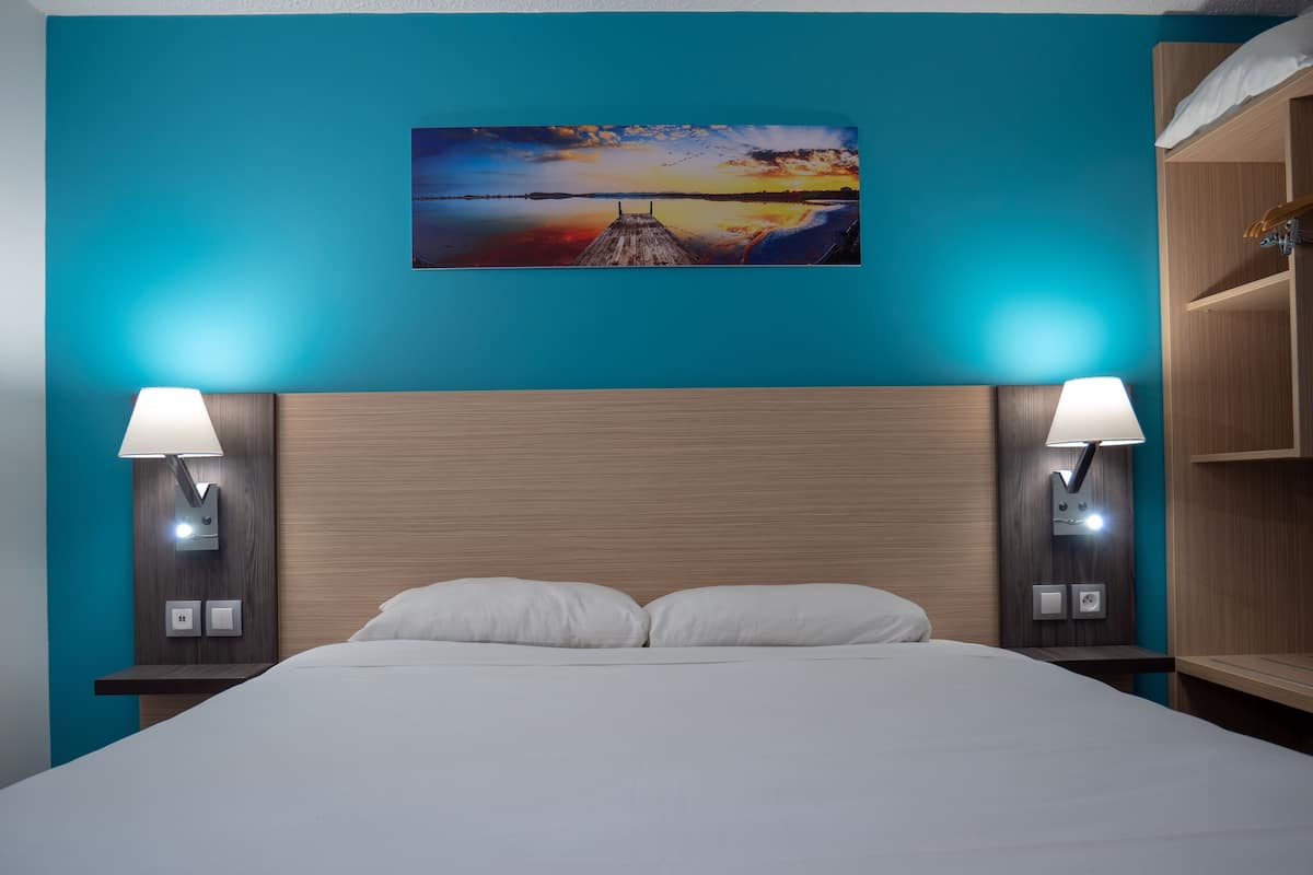 A hotel room with a large bed, two bedside tables with lamps, a wooden headboard, and a painting of a sunset over water on the wall.
