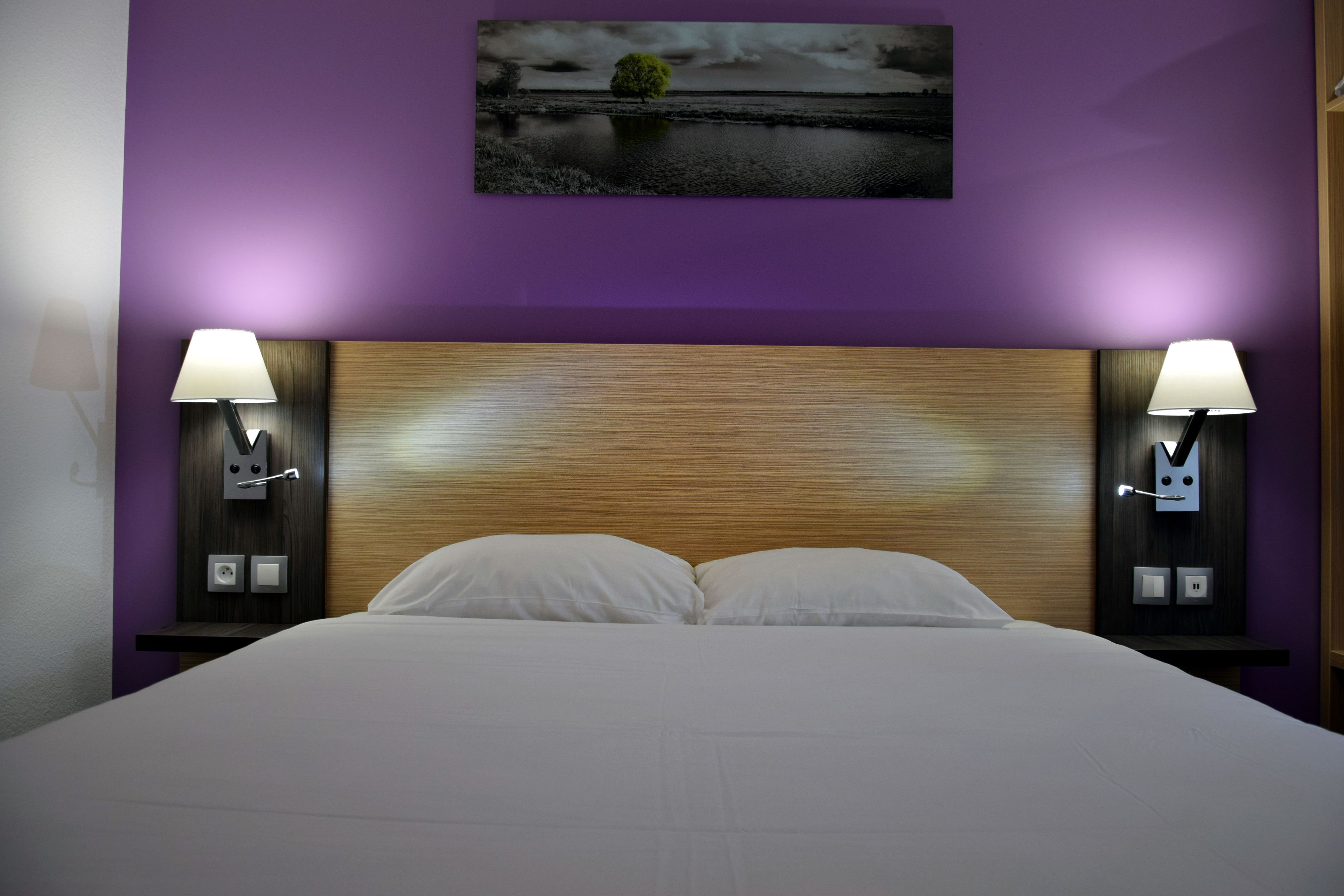 A hotel room with a large bed, two bedside tables with lamps, a wooden headboard, and a wall-mounted picture.
