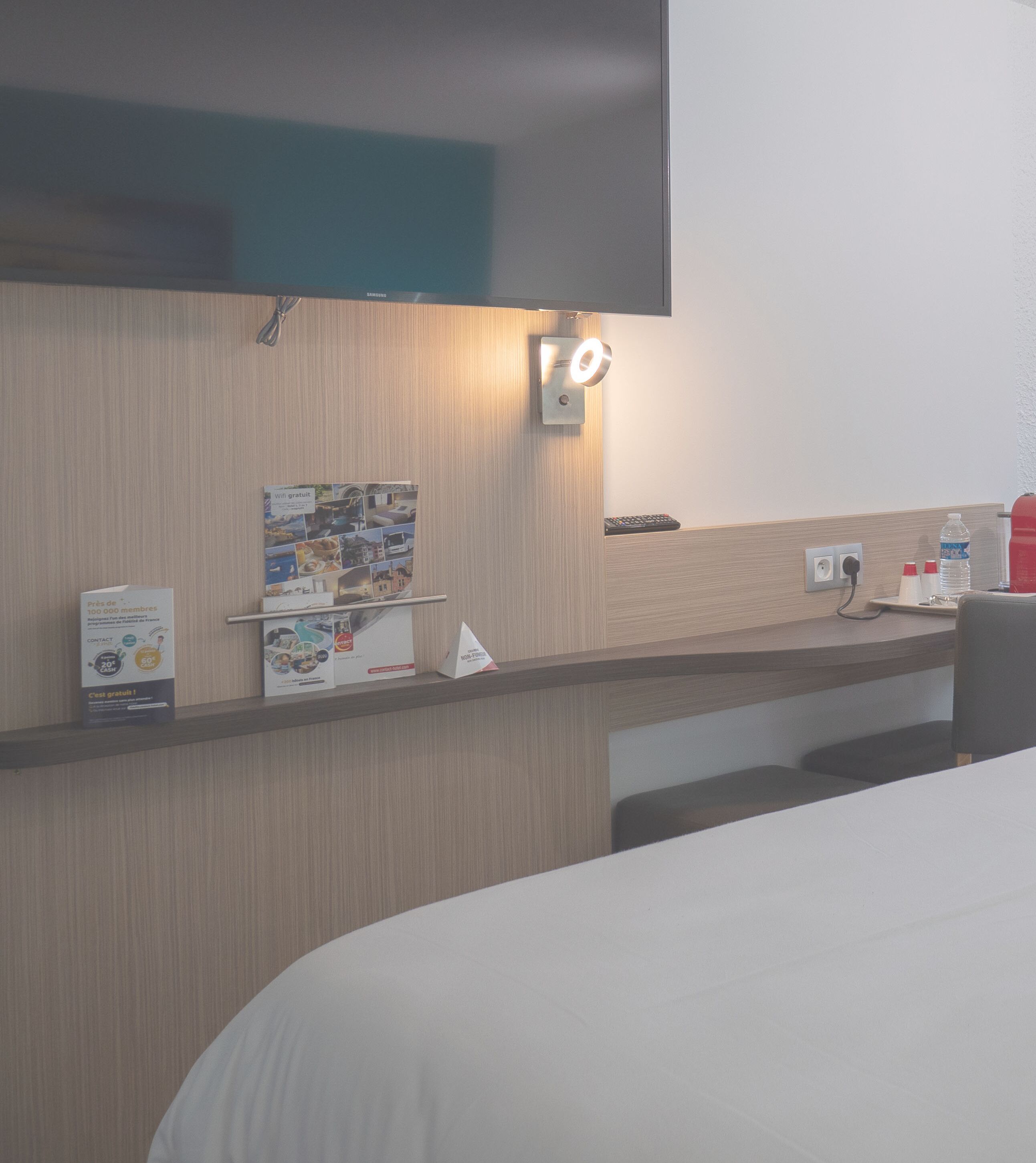A hotel room with a bed, a flat-screen TV mounted on the wall, a small table with a remote control, a water bottle, and a wall-mounted light.
