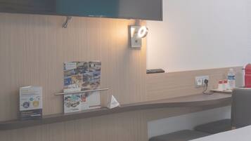 A hotel room with a bed, a flat-screen TV mounted on the wall, a small table with a remote control, a water bottle, and a wall-mounted light.