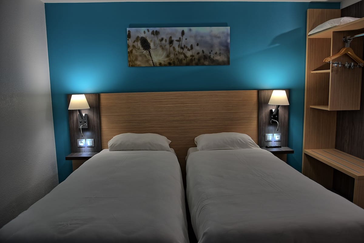 A hotel room with two beds, wooden headboards, bedside lamps, a painting on the wall, and a wooden wardrobe.