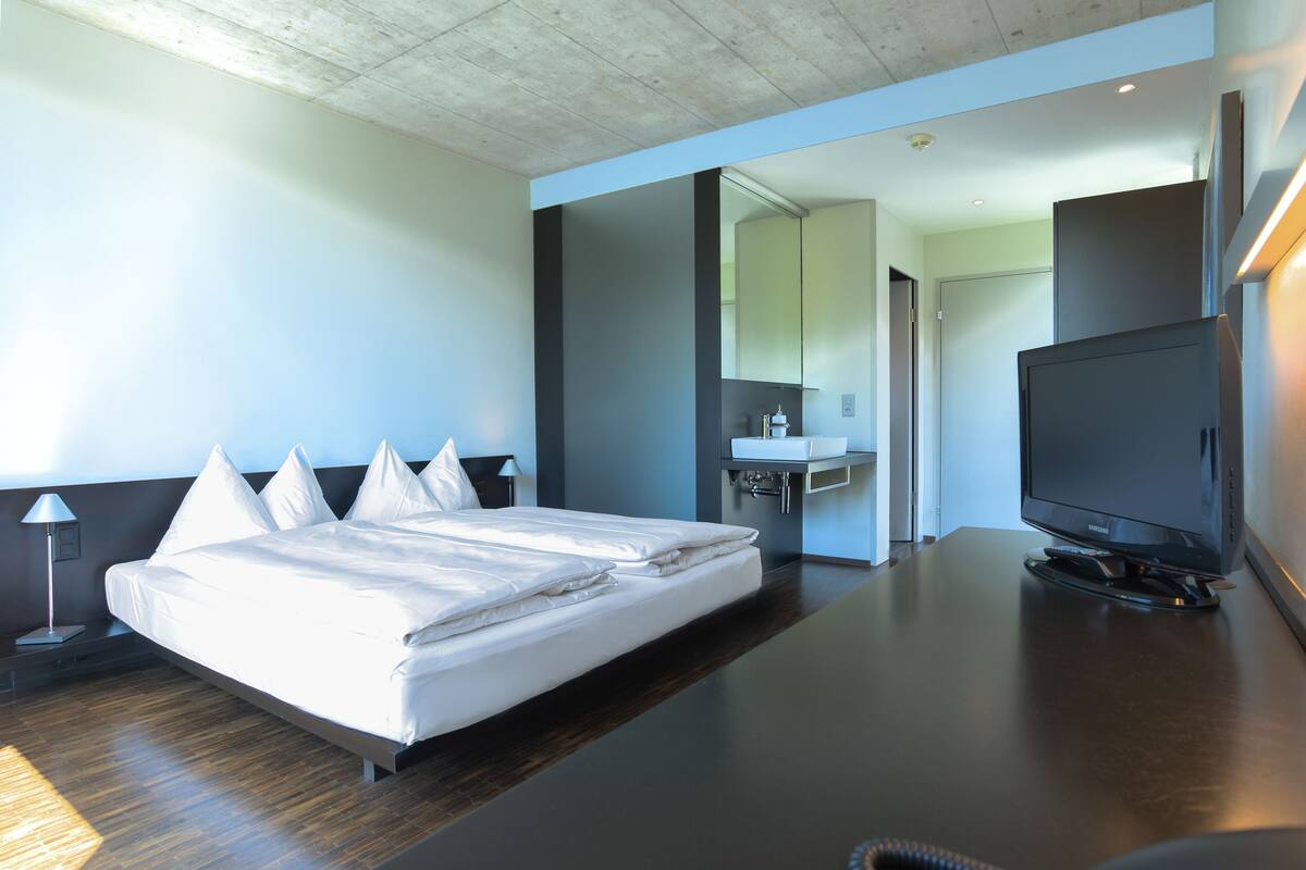 Ein modernes Hotelzimmer mit einem großen Bett, einem Flachbildfernseher, einem Schreibtisch und einem durch eine geöffnete Tür einsehbaren Badezimmer.