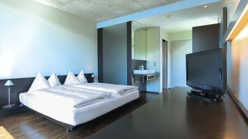 Quarto de hotel moderno com uma cama grande, televisão de tela plana, escrivaninha e banheiro visível através de uma porta aberta.