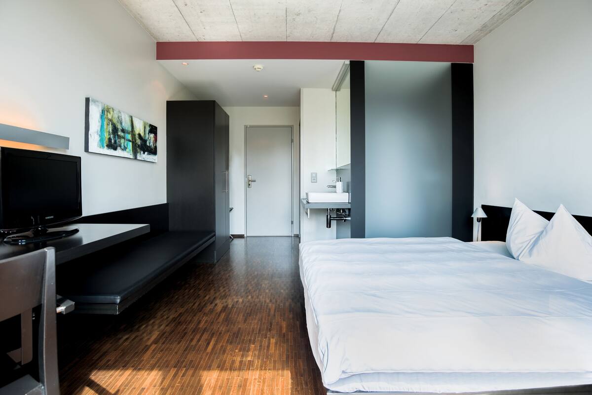 Ein modernes Hotelzimmer mit Bett, Schreibtisch mit Fernseher, Bad mit Waschbecken und Spiegel sowie einer Tür zu einem weiteren Raum.