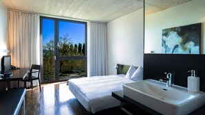 A modern hotel room with a large bed, a desk with a computer, a bathroom with a sink and mirror, and a view of trees through the window. - Hotel Kreuzlingen am Hafen (Kreuzlingen)