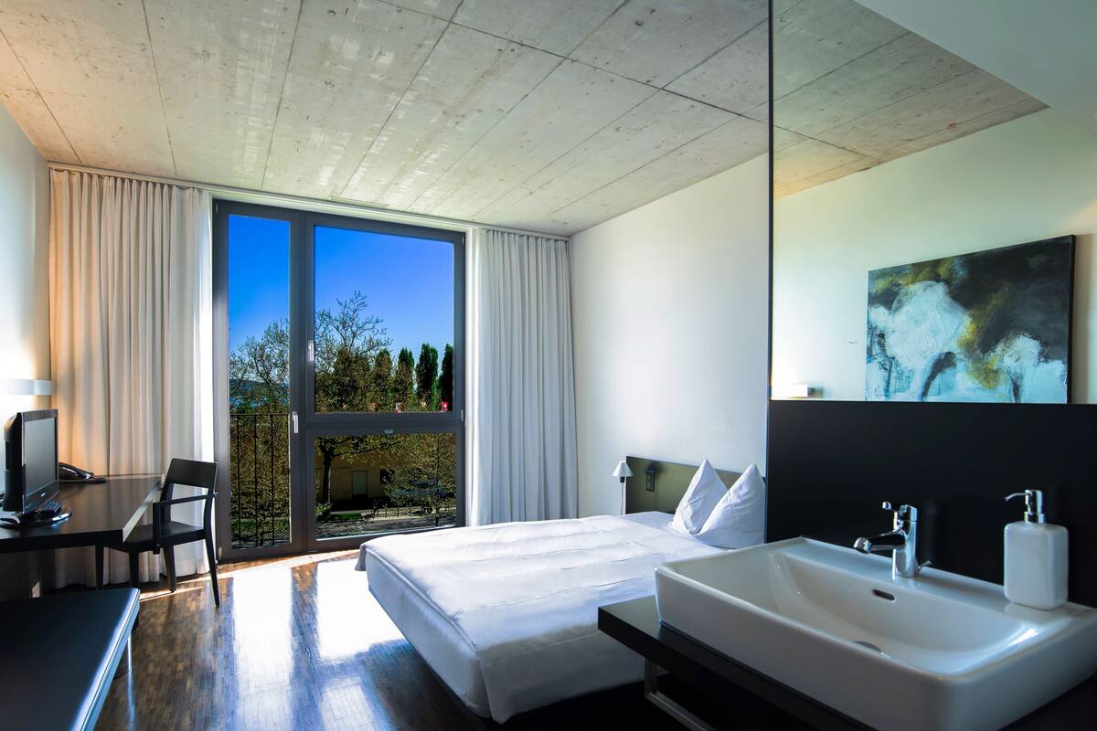 Ein modernes Hotelzimmer mit einem großen Bett, einem Schreibtisch mit Computer, einem Badezimmer mit Waschbecken und Spiegel sowie Blick auf Bäume durch das Fenster.