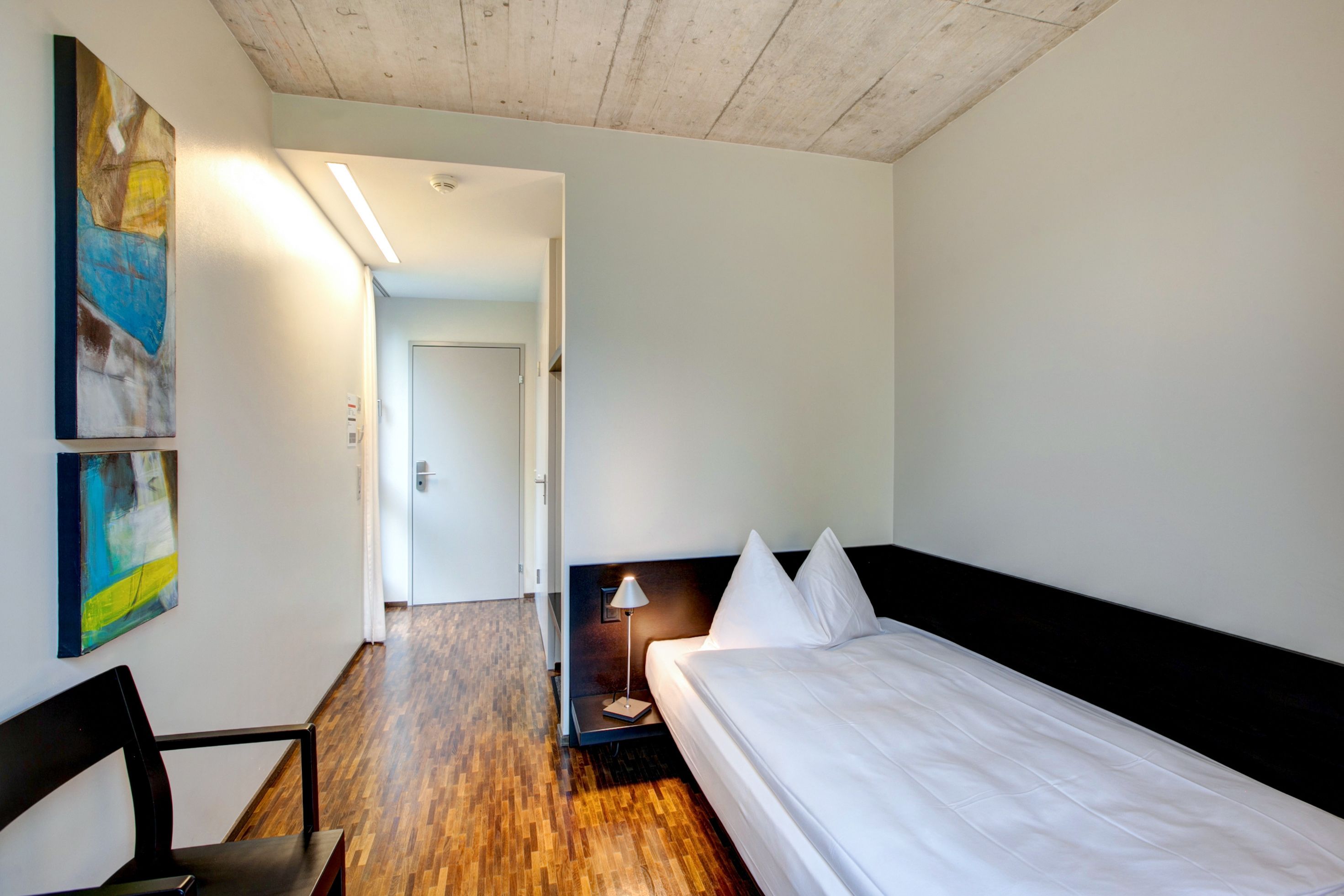 Een hotelkamer met een houten vloer, een bed met witte lakens, een bureau met een stoel en een schilderij aan de muur.