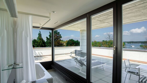 A modern bathroom with a bathtub, a glass shower enclosure, and a large window overlooking a patio area with outdoor furniture. - Hotel Kreuzlingen am Hafen (Kreuzlingen)