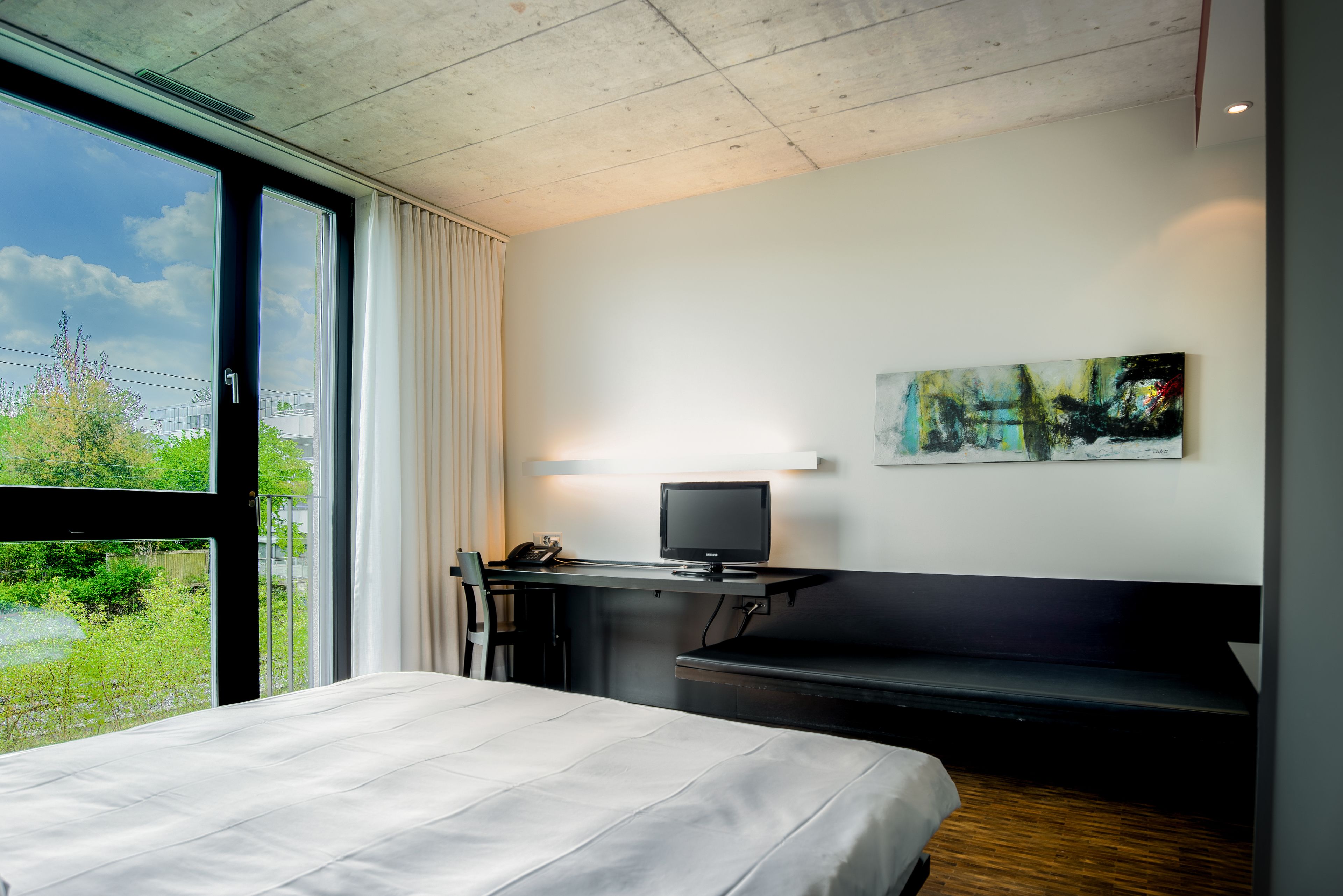A hotel room with a bed, a desk, a chair, a television, and a painting on the wall.