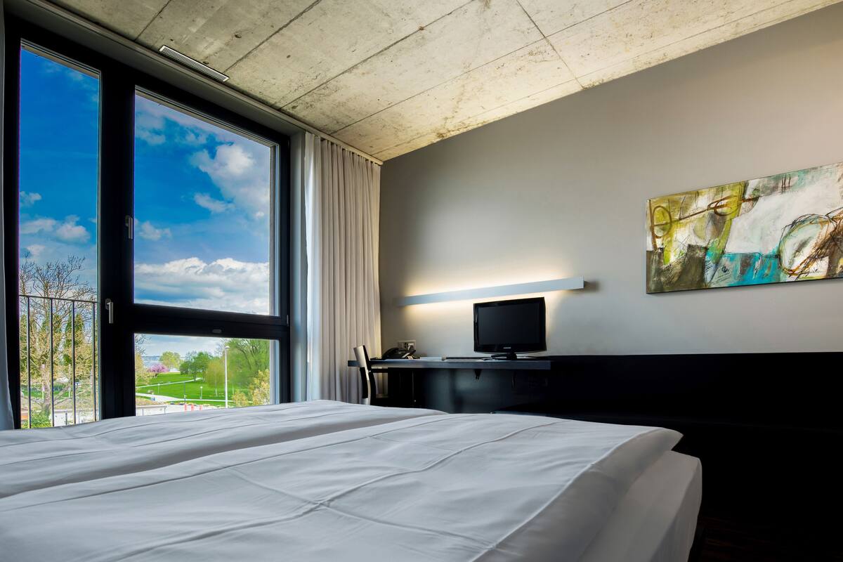 Ein Hotelzimmer mit einem großen Bett, einem Schreibtisch mit Computer, einem Fernseher und einem Bild an der Wand.