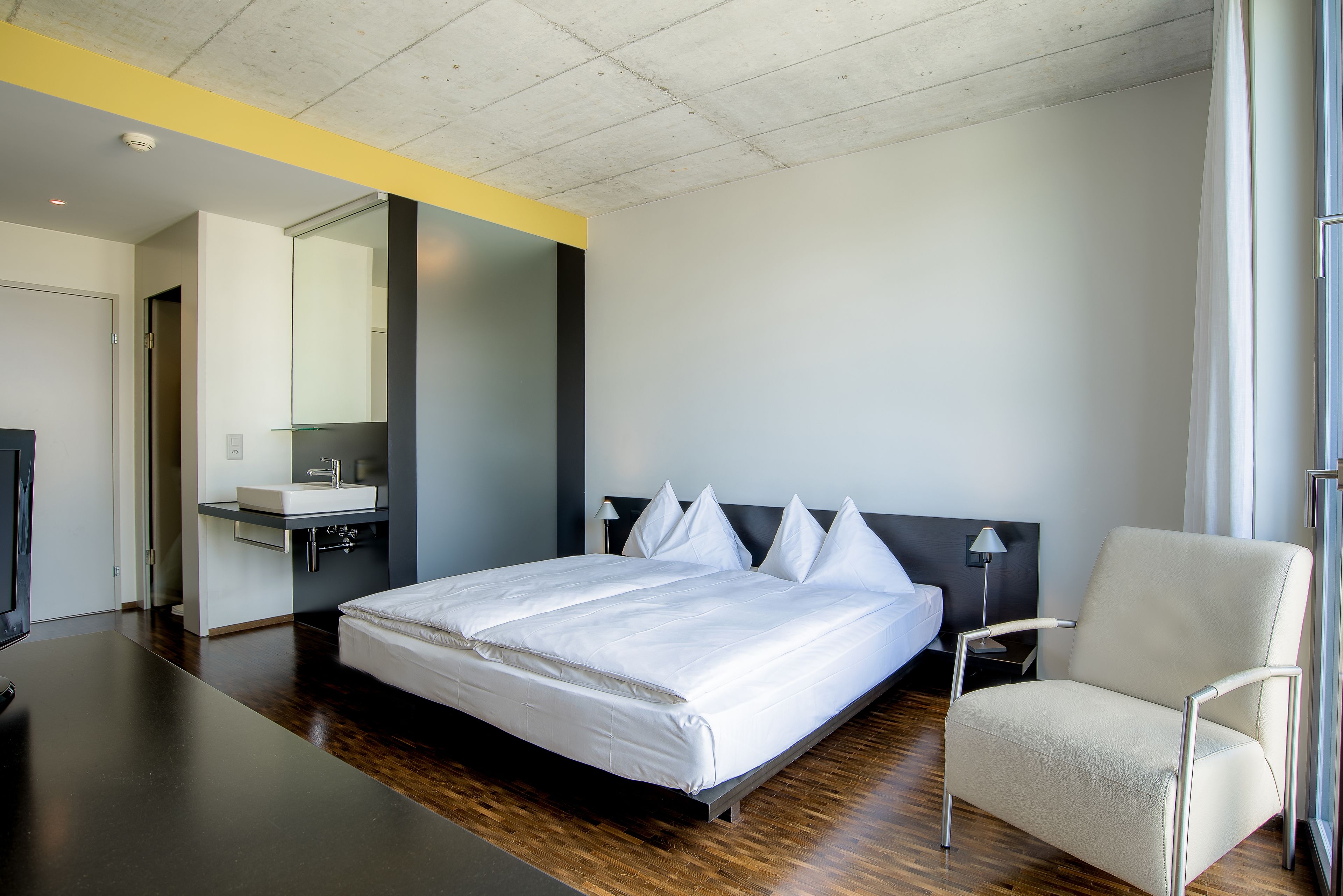 A modern hotel room with a large bed, a bedside table, a chair, a desk, a mirror, and a window.