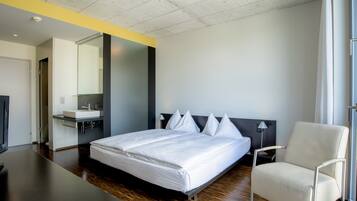 A modern hotel room with a large bed, a bedside table, a chair, a desk, a mirror, and a window.