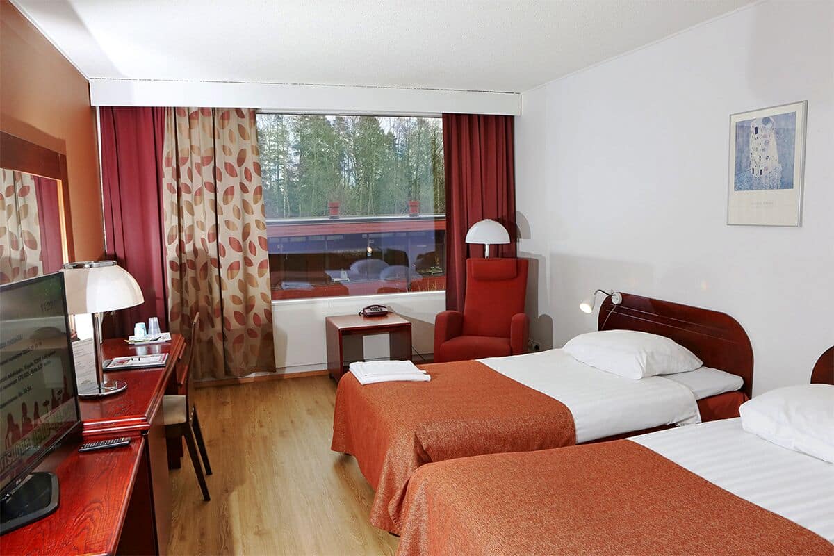 Une chambre d’hôtel avec deux lits, un bureau avec un ordinateur, une chaise, une petite table et une fenêtre avec des rideaux.