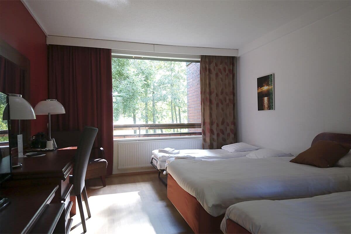 Une chambre d’hôtel avec deux lits, un bureau avec une chaise, une fenêtre avec des rideaux et un radiateur.