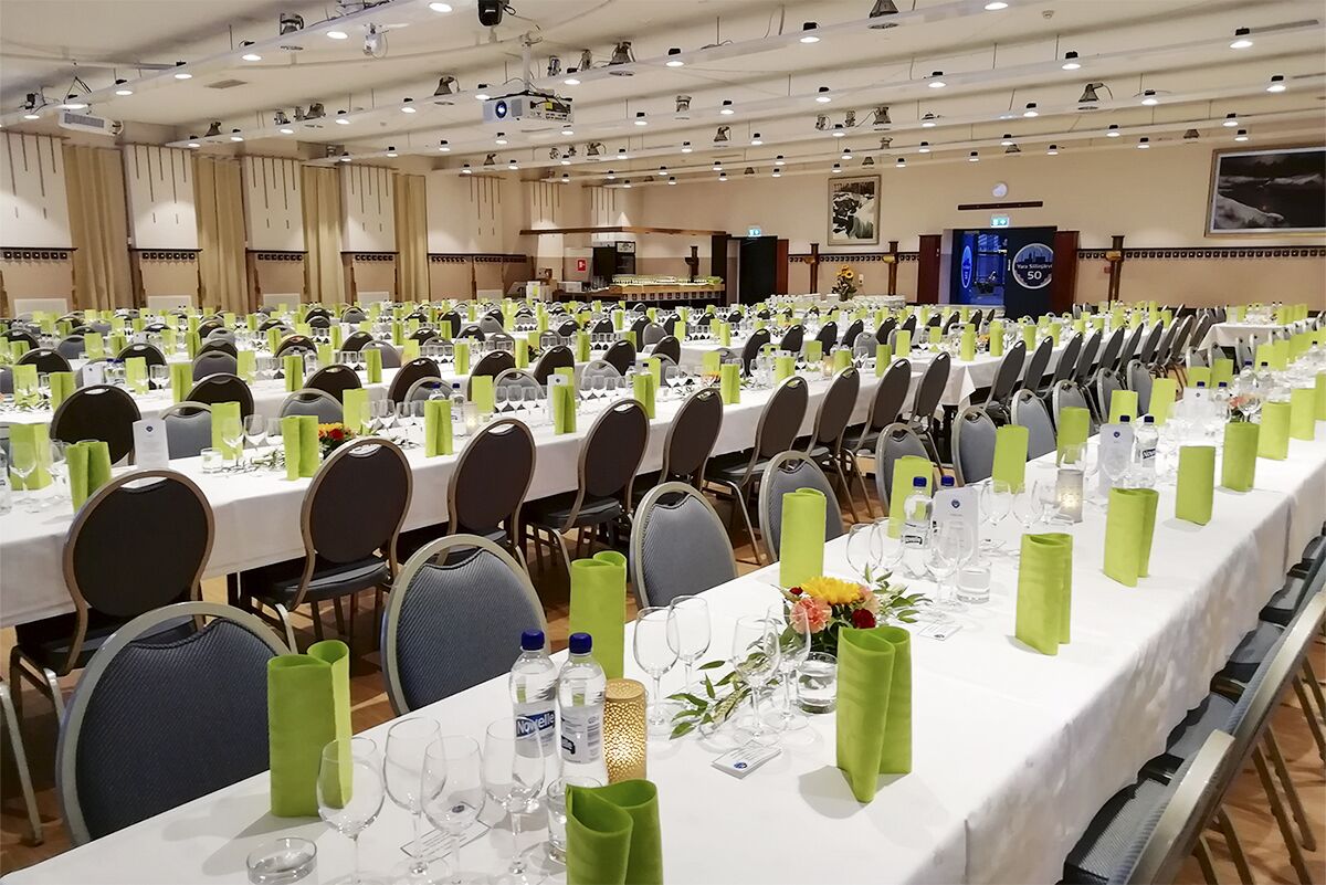 Un salón de conferencias con filas de mesas dispuestas para una reunión, equipado con sillas y cubiertas con manteles verdes.
