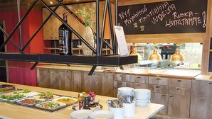 Montaje de bufé con diversas opciones de comida y platos apilados, una pizarra con los platillos del menú, y equipo de cocina al fondo.