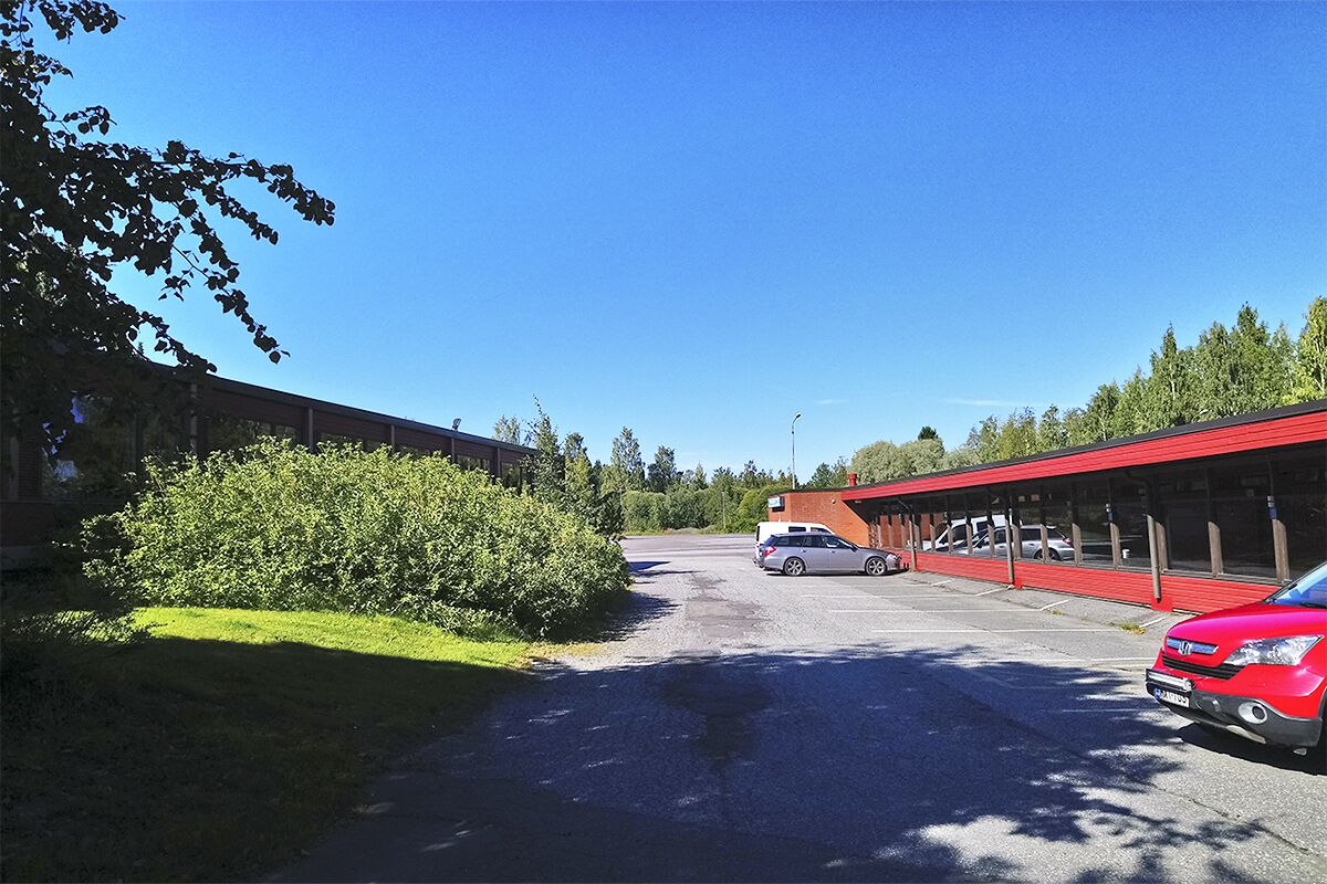 A parking area with a red car and a white car parked. There is a building with a red roof and a covered area.