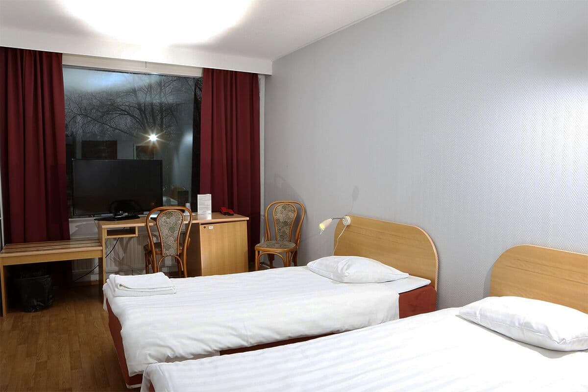 Une chambre d’hôtel avec deux lits simples, un bureau avec une chaise, une armoire et une grande fenêtre avec des rideaux.