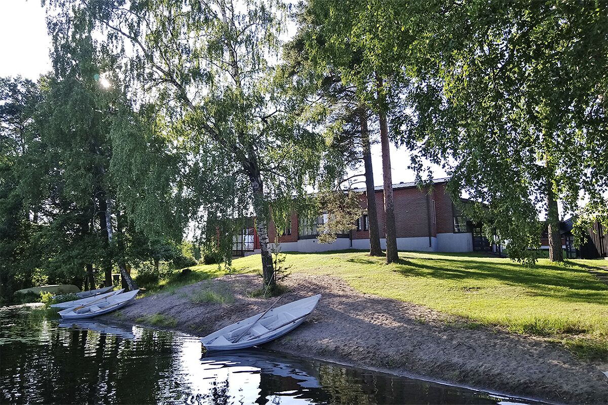 Joenrantavyöhyke, jossa on laivoja laiturissa, puita ja taustalla näkyvä rakennus.