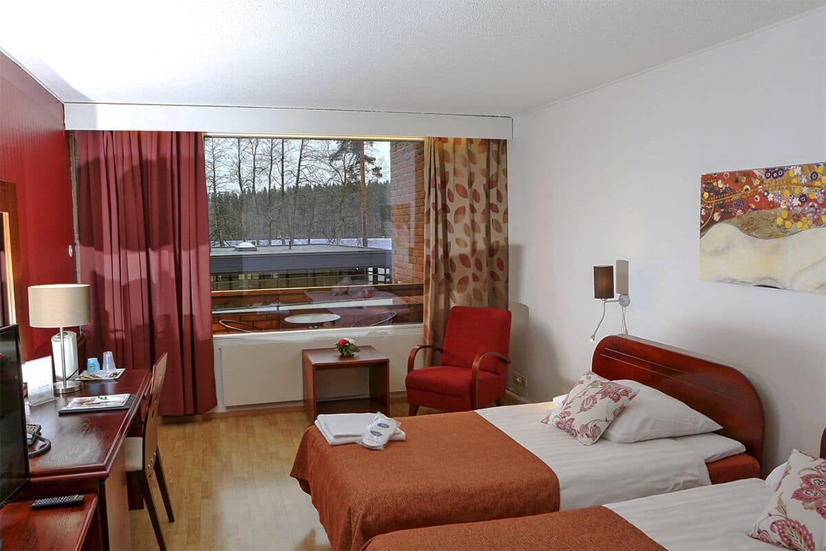 Une chambre d’hôtel avec deux lits, un bureau avec une chaise, une télévision, une fenêtre avec des rideaux et un tableau au mur.