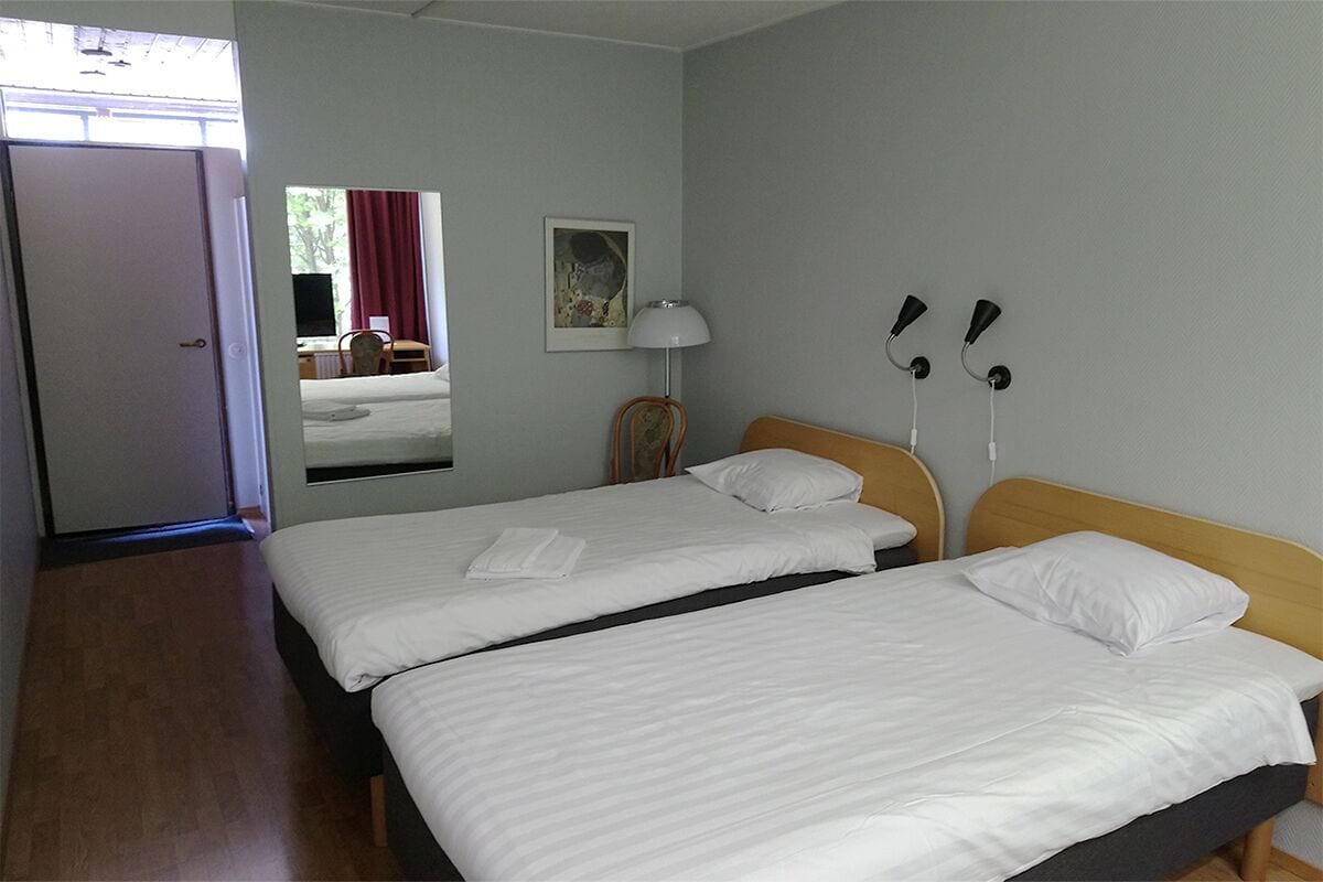 A hotel room with two single beds, a wooden headboard, a desk with a chair, a window with a red curtain, and a wall-mounted lamp.