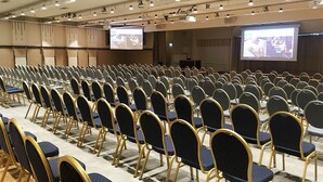 Un salón de conferencias con filas de sillas y una pantalla grande para presentaciones.